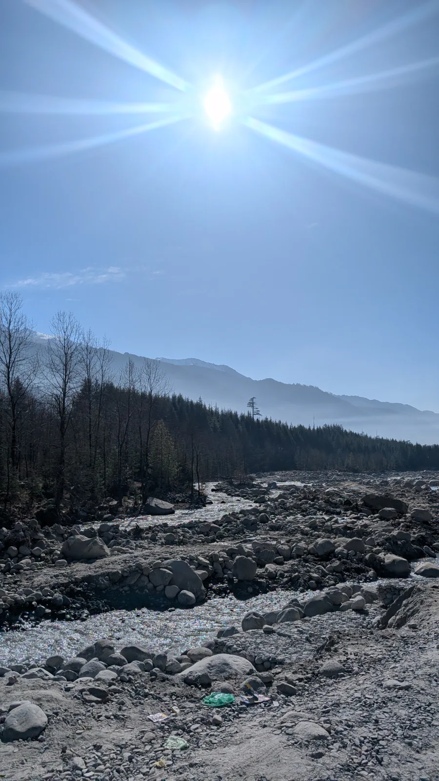 Sunburst over a rocky mountain riverbed, with a pine forest and hazy ridges behind